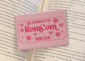 Open book with a pink magnet reading 'A Member of the RomCom Book Club' on a hardback fridge magnet.